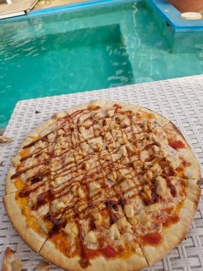 The image captures a close-up, angled view of a large, freshly baked pizza positioned next to a swimming pool. The pizza, which appears to be approximately 12-14 inches in diameter, is cut into multiple slices and takes up a significant portion of the right side of the frame. Its crust is golden brown, and the surface is generously topped with melted white cheese, chunks of cooked chicken, and a noticeable red base sauce (likely tomato sauce). A dark, glossy sauce, possibly barbecue sauce, is artfully drizzled in a crisscross pattern over the entire pizza, adding visual appeal and contrast. A few small, scattered crumbs or pieces of crust are visible on the surface beneath the pizza.

The pizza rests on a light-colored, possibly white or off-white, woven surface, which resembles outdoor furniture such as a patio table or chair. To the left of this surface and the pizza, a swimming pool or a similar body of water is prominently featured. The water is a vibrant turquoise or teal color, suggesting it is clear and clean. Ripples and reflections are visible on its surface, indicating movement or light playing on the water. A blue-colored edge or rim of the pool is visible along the top and left side, contrasting with the water's color. The overall setting suggests an outdoor, leisurely environment, perhaps during daytime given the natural light. No people are clearly visible, nor is any discernible text present in the image.