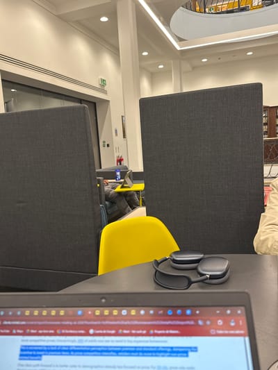 The image shows an interior space, likely a library or study area, in Hull, United Kingdom. The foreground features a laptop displaying text, with headphones resting on a gray partition next to a yellow chair. Other people are visible in the background, seated and engaged in study activities. The environment includes modern furniture, gray dividers, and a prominent window.