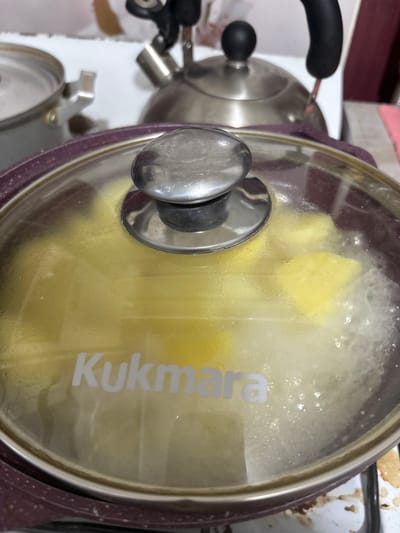 The image shows a close-up of a pot with a clear glass lid. Inside the pot, yellow food items, possibly potatoes, are being cooked in boiling water, creating steam and bubbles visible through the lid. The brand "Kukmara" is visible in white lettering on the lid. In the background, a stainless steel kettle with a black handle is partially visible, along with another pot on the left. The scene appears to be a kitchen counter. The image does not provide any information about the city of Княгинино, Russia.