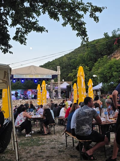 The image captures an outdoor event, likely a music festival, set in Gara Oreshets, Bulgaria. The scene is oriented vertically, with the sky and trees on the left and the event setup on the right. A stage with a band performing is visible under a large white tent, adorned with stage lights. Yellow umbrellas are spread out, providing shade for attendees seated at tables.

People are gathered, sitting at tables and chairs, watching the performance. Some are facing the stage, while others are seen from the back. The overall atmosphere appears relaxed and engaged, with individuals enjoying the event. The time of day seems to be late afternoon or early evening, as indicated by the soft light and the presence of the moon in the sky. The weather appears to be pleasant, suitable for an outdoor gathering.

Behind the stage, a lush green hillside covered with trees dominates the background, suggesting a natural or semi-rural setting. The ground appears to be a mix of dirt and grass.

Visible text on the tent above the stage reads in Cyrillic script: "ФОЛКЛОРЕН ФЕСТИВАЛ 'НИВА ВОДА' ВЕНЕЦА". This translates to "Folklore Festival 'Niva Voda' Venetsa". This text clearly identifies the nature and name of the event.