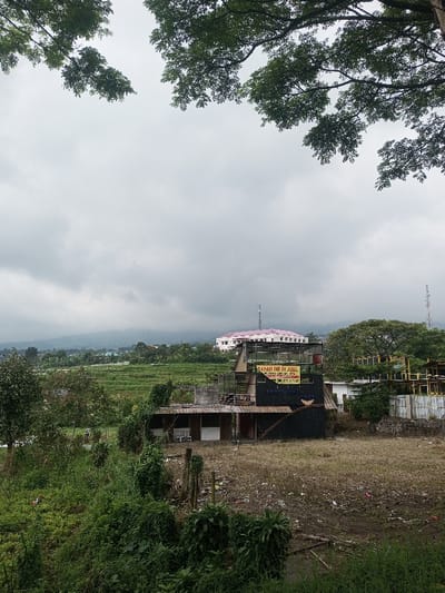 The image displays an outdoor scene in Lunugamwehera, Sri Lanka, under an overcast sky. The upper foreground is partially obscured by dark green tree foliage. The ground in the immediate foreground consists of a patch of dry, light brown grass and scattered debris on the right, and dense, unkempt green vegetation on the left, with visible wooden posts.

In the midground, a multi-story structure dominates, featuring a lower section constructed from dark, possibly corrugated metal, and an upper section with an open framework. A yellow banner is affixed to the upper section, bearing the text "TANAH INI DI JUAL". To the right of this structure, a smaller white building is partially visible, followed by a larger, geometrically structured yellow object, possibly another building or decorative installation.

Further in the background, green terraced fields, likely agricultural, extend horizontally across the frame. Above these fields, a prominent light-colored building with a distinctive pink or red roof stands out. A tall, thin communication tower is visible to the right of this building. The horizon is marked by distant, hazy mountains under the uniformly grey, cloudy sky. No individuals or active interactions are present in the scene.