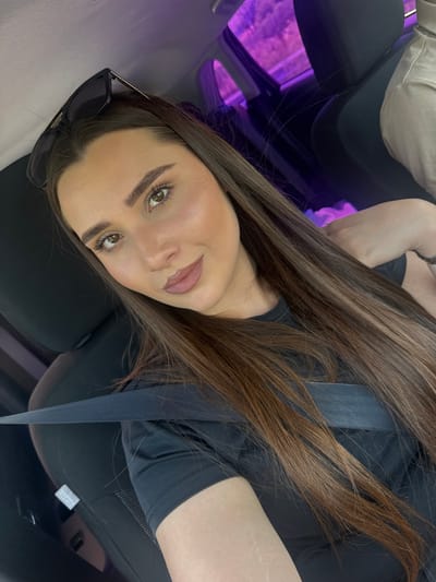 A young woman with long, dark brown hair is taking a selfie in a car. She is wearing a black t-shirt and sunglasses are perched on top of her head. Her seatbelt is fastened across her chest. The interior of the car is dark, with purple lighting visible in the background, possibly from interior lights or reflections. A portion of another person's shoulder and arm, wearing a light-colored shirt, is visible in the upper right corner of the frame. The scene appears to be within a car, potentially in or near the city of Razliv, Bulgaria, though this location is not explicitly depicted in the image itself.