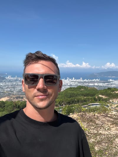 A medium shot of a man wearing sunglasses and a black t-shirt is oriented vertically. The backdrop consists of a city with numerous white buildings situated along a coastline with a body of water, possibly the ocean or a bay. There are patches of green vegetation, likely hills or mountains, surrounding the city. The sky is blue with scattered white clouds. The location may be Diên Khánh, Vietnam.