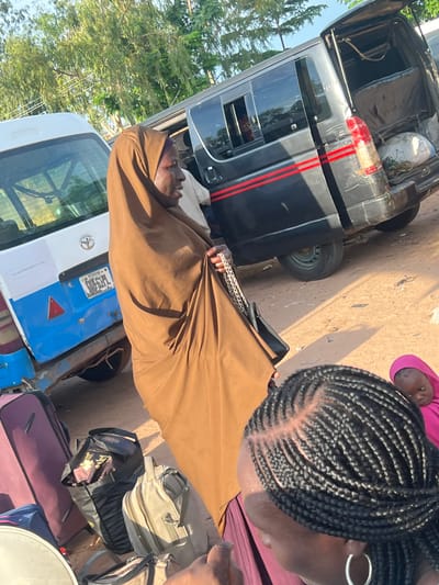 The image depicts an outdoor scene in Gbaza, Plateau State, Nigeria, centered around two vans and several individuals.

In the center, a woman wearing a light brown full-body covering (possibly a jilbab or khimar) and matching head covering stands facing right, holding a black handbag with a silver chain strap. Her face is visible in profile. To her left, the rear of a white and blue Toyota van is visible, featuring a Nigerian license plate that reads "PLATEAU Gbaza." A red triangular reflector is affixed to its lower rear.

To the right of the woman, a dark grey or black van with two parallel red stripes on its side is parked with its rear hatch open, revealing a cargo area containing a white sack and a dark fabric cover. The ground is unpaved, appearing sandy or dusty brown, with shadows indicating bright daylight.

In the immediate foreground, the back of a person's head with black braided hair and a silver hoop earring on the right ear is visible, wearing a purple garment. To the right of this person, a young child wearing a pink head covering is partially visible, looking downwards.

Several pieces of luggage, including a purple wheeled suitcase, a black soft-sided bag, and a lighter-colored backpack, are on the ground in the lower-left section of the frame. Green trees are visible in the background, along with faint overhead wires in the upper left. A light-skinned individual in a white shirt is partially visible between the woman in brown and the dark grey van.