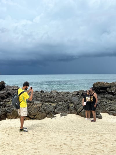 A man in a yellow shirt and khaki shorts stands on a sandy beach in Phru Nai, Thailand, holding a smartphone to take a picture of a couple standing on rocky terrain. The couple, a man in a black tank top and a woman in a black t-shirt and skirt, pose with their arms around each other. The ocean is visible in the background under a cloudy sky. Rocks and pebbles cover the area between the sand and the sea.