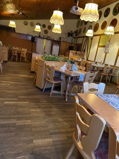 The image depicts the interior of a vacant restaurant or gaststätte located in Eppelheim, Germany. The scene is illuminated by warm light primarily from multiple ornate, cream-colored fabric pendant lamps with fringed edges suspended from a slanted wooden plank ceiling. A dark-bladed ceiling fan and a white ceiling-mounted projector are also visible overhead.

The flooring consists of dark brown faux wood planks arranged in a staggered pattern. The room is furnished with numerous light-colored wooden tables and matching chairs. The chairs feature rounded backrests and maroon fabric seat cushions with an understated pattern. Many tables are set with white folded napkins, small decorative plants in brown bottles, and table runners displaying a light-colored pattern, possibly an olive branch motif.

A long, light-toned wooden counter with integrated planters containing green foliage separates dining areas in the mid-ground. The walls exhibit distinct styles: the left wall is a solid tan with a dark horizontal beam, while the right wall features a traditional half-timbered aesthetic with light yellow plaster infill and dark brown beams. Both walls are adorned with numerous decorative circular plates. In the background, a black PA speaker on a stand is visible. Shelves behind the counter hold various decorative objects including small statues and trophies. No individuals are present within the frame.