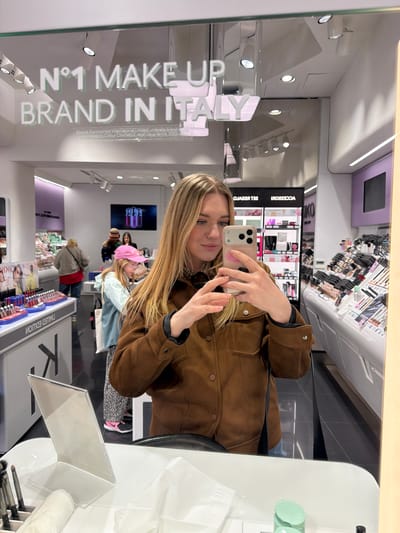 A young woman is taking a selfie in a mirror, positioned in a brightly lit retail setting. The mirror's reflection captures the interior of the store, displaying makeup displays and other shoppers. The woman is wearing a brown jacket. A prominent sign stating "N°1 MAKE UP BRAND IN ITALY" is visible. The location is a retail store, possibly in the city of Siena, Italy.
