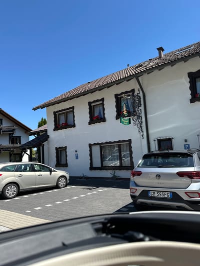 In the city of Schönwald im Schwarzwald, Germany, a bright white, two-story building with a brown-tiled roof stands under a clear blue sky. The building features dark-framed windows with decorative red flowers in window boxes. A green sign with the words "Schwarzwald Tanne" hangs from a black, wrought-iron bracket on the building's facade, indicating it is likely a business, possibly an inn or restaurant. Two cars are parked in front of the building on a paved area with white parking lines. On the left, a silver-gray station wagon is parked at an angle. On the right, a silver-gray SUV is parked, facing away from the viewer, with its license plate visible. The scene appears to be a sunny day in a residential or commercial area.