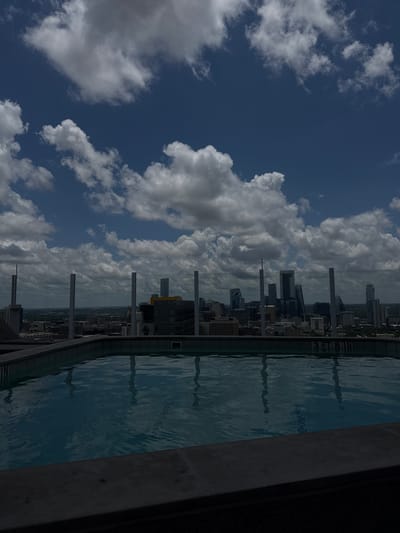 An elevated, static long shot captures a rooftop swimming pool overlooking the Austin, Texas, USA, downtown skyline under a partly cloudy sky. The foreground features the dark grey coping of the infinity-edge pool, with the pool's dark blue water occupying the lower portion of the frame. Reflections of the sky and distant buildings are subtly visible on the water's surface. On the far side of the pool, submerged white tiles reveal the depth marker "3 FT 8 IN" in black lettering. A transparent glass railing, supported by vertical metal posts, extends across the midground, separating the pool from the cityscape. Beyond the railing, the Austin skyline is prominent, featuring numerous high-rise buildings and skyscrapers under diffused daylight. Notable structures identifiable include the distinct 'Jenga'-like structure of The Independent on the right side of the main cluster, and the sleek design of the Austonian towards the center-left. The background consists of a deep blue sky dotted with numerous bright white and grey cumulus clouds, indicating partial cloud cover. No individuals are present in the scene, and no specific actions or interactions are depicted.