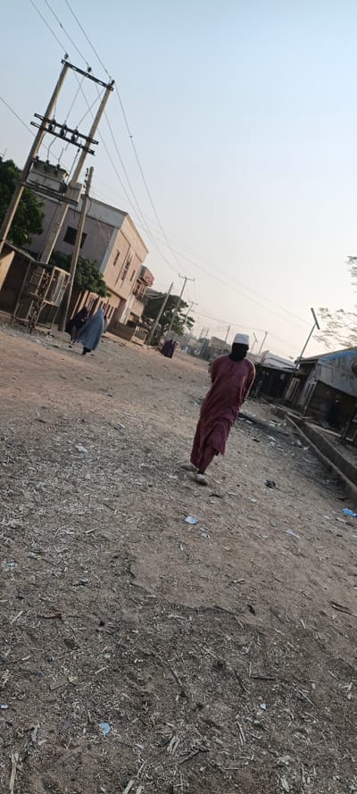 A street scene shows a man wearing a maroon robe and white cap walking towards the viewer. Another person in a full blue robe walks in the opposite direction. The unpaved street is filled with debris. Buildings line both sides of the road. Power lines are visible above. The sky is a light blue.