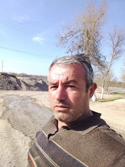 A male subject, approximately 40-50 years of age, with short, greying dark hair and a short greying beard/stubble, is depicted in an outdoor, eye-level, selfie-style photograph. He has a tan complexion, a neutral to serious expression with a slight furrowed brow, and is looking directly at the camera. He wears a collared polo-style shirt, brown in color with a textured fabric, featuring a diagonal stripe pattern across the upper left chest area in shades of blue, brown, red, and beige. Strong sunlight illuminates the subject from his left, casting distinct facial shadows.
The scene is located in Yangi Chinoz, Uzbekistan. The immediate foreground and mid-ground consist of dry, sandy earth with visible vehicle tire tracks. A small wet area or puddle is present within the tracks to the subject's left. In the left background, large, uneven mounds of earth and gravel are observed, indicative of an excavation or construction site. The right background features several bare deciduous trees, one with a white-painted lower trunk, leading to what appears to be a body of water or marshy area in the far background. Power lines are visible in the upper left quadrant of the sky, connected to a utility pole partially obscured on the far left. The sky is bright blue with minimal cloud cover.