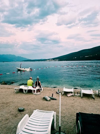 This image depicts a tranquil scene at a beach in Đenovići, Montenegro, during what appears to be late afternoon or early evening. The sky is filled with dramatic, textured clouds, a mix of soft pinks and muted blues, with a small, pale moon visible. The sea is a deep turquoise, with gentle ripples. In the background, a chain of white and red buoys demarcates a swimming area. A small white boat with a dark outboard motor is moored in the water. Along the shoreline, there are several white plastic lounge chairs, some occupied and some empty.
In the foreground, two individuals are seated on a lounge chair facing the water. One, a male with grey hair, wears a bright green t-shirt. Beside him, a person with dark hair wears a dark top. Another lounge chair is positioned in front of them. Further to the right, two more white lounge chairs are empty, one with a folded pink towel on it. The beach itself is covered in small pebbles and rocks. In the mid-ground, near the water's edge, are more white lounge chairs and scattered rocks. A dark object with wheels, possibly a storage container or equipment, is visible in the lower right corner of the frame. The overall atmosphere is peaceful and relaxed, suggesting a quiet end to a day at the beach.