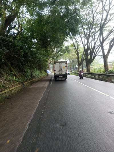 The image depicts an outdoor scene on a two-lane asphalt road in the city of YonPhoogla, Bhutan, under overcast conditions. The road surface is wet, suggesting recent precipitation.
In the foreground, the perspective is from a moving vehicle, facing forward. A solid white line delineates the left edge of the road, separating it from a paved shoulder or path. A stone retaining wall is visible along the base of a heavily vegetated embankment on the far left. Large trees with dense green foliage line this embankment, overhanging the road.
In the midground, a white box truck with two yellow triangular hazard symbols on its rear doors is positioned in the left lane, moving away from the camera. Its license plate, partially visible, appears to be a white plate with black characters. To the right of the truck, a person wearing a white helmet and a reddish-brown jacket is operating a white scooter in the right lane, also moving away. A solid yellow line separates the two lanes.
Further down the road, in the background, a white car, possibly a sedan, is parked or stopped on the paved shoulder to the left of the truck. Another white car, possibly an SUV, is visible in the right lane beyond the scooter. A metal guardrail runs along the right side of the road, and beyond it, additional trees with sparser foliage and distant hills are visible. The sky is uniformly light and overcast.