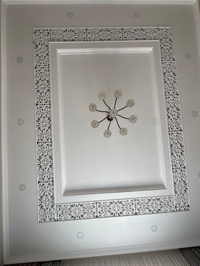 This media file showcases an interior ceiling with a decorative recessed design. The primary focus is a white, multi-tiered ceiling featuring ornate white decorative molding around a central rectangular area. Within this central rectangle, a modern chandelier with multiple clear glass globes on dark metal arms hangs from the ceiling. The surrounding ceiling area has recessed circular light fixtures. The decorative molding itself is intricate, with a repeating pattern of floral and scrollwork elements. The lighting suggests an indoor setting, likely during daylight hours due to the ambient light. The overall impression is one of elegant interior design. There is no visible text. The location context is Taraz, Kazakhstan.