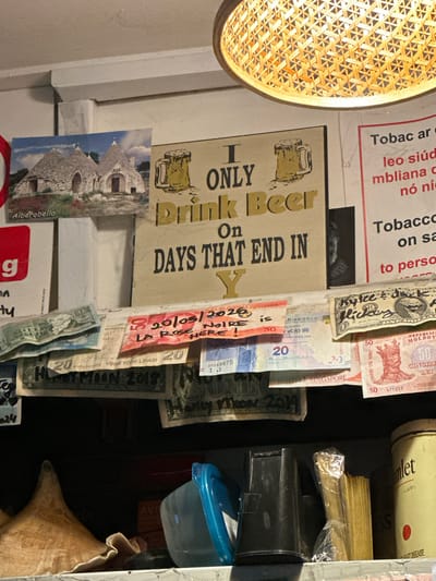 The image shows a cluttered shelf with various items. A framed sign with the words "I ONLY Drink Beer On DAYS THAT END IN Y" is prominently displayed. Below the sign, several pieces of currency and what appear to be receipts or notes are taped or pinned to the shelf. These include what look like US dollars, Turkish Lira, and Hong Kong Dollars. There is also a postcard of Alberobello, Italy, visible on the left. To the right of the sign, a poster with text in an unknown language, possibly Irish given the mention of Westport, Ireland, is partially visible. In the foreground, there are various objects, including a blue plastic container, some black plastic items, and a stack of what look like wooden stirrers or skewers. A metal tin with the word "Hamlet" on it is on the far right. The overall impression is of a casual, perhaps slightly untidy, personal space. There are no visible people in the image.