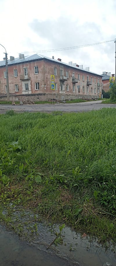 The image shows a long, dilapidated, salmon-colored apartment building with multiple stories and balconies. The building appears to be in a state of disrepair, with peeling paint and visible signs of age. In front of the building, there is a paved area that looks like a road or intersection, and a patch of lush green grass in the foreground. There are several traffic signs visible, including a yellow diamond-shaped sign with a pedestrian crossing symbol and another sign depicting children walking. The sky is overcast. The apartment building is identified as being in the city of Lysva, Russia, specifically at the intersection of Lenina Street and Komsomolskaya Street. This intersection is known for its proximity to the former Lysva Metallurgical Plant, which contributes to the industrial and somewhat run-down character of the area. The overall scene depicts a somewhat bleak urban environment with evidence of both residential and industrial past.