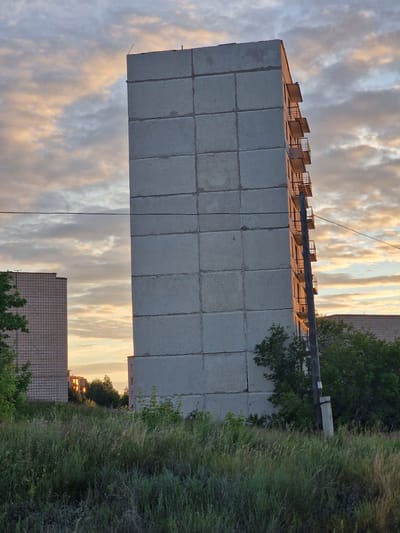 An image captures a concrete apartment building in Kashgar City, China, during what appears to be sunset. The sky is filled with a mix of clouds, some tinged with orange and yellow from the setting sun, while others are a pale blue and gray. The building itself is tall and rectangular, constructed from large, square concrete panels. Balconies with rusty metal railings jut out from one side of the building, spaced evenly. A weathered wooden utility pole stands near the building, supporting a single black wire that stretches across the frame. In the foreground, tall, dry grass and weeds create a wild, overgrown field. To the left, another apartment building with a grid-like facade is visible, and to the right, more trees and another building can be seen in the distance. The overall scene is somewhat stark, with the industrial architecture contrasted by the natural elements and the soft, atmospheric light of the sky. There are no people visible in the image.