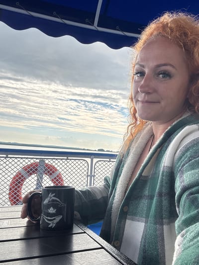 A woman with curly red hair and blue eyes is sitting at a table on a boat, holding a black mug with whale and paper boat designs. She is wearing a green and white plaid jacket over a white fuzzy-lined shirt. The boat has a blue canopy and a life preserver is visible in the background. The sky is cloudy. The image is taken outdoors with a slight angle.