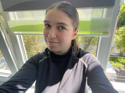 A young person with light skin and short gray hair is looking directly at the camera. They are wearing a dark athletic top with white stitching along the sleeves. The photo is taken at an upside-down angle. Behind the person is a window with roller shades, colored gray, white, and green. Outside the window are trees with green leaves. On the left side of the image, a sign with Cyrillic text is visible, reading "ДОСТАВКА ВОДЫ." Based on the text on the sign, the location is likely Ukraine. There is no other information about the city of Cherliany available.