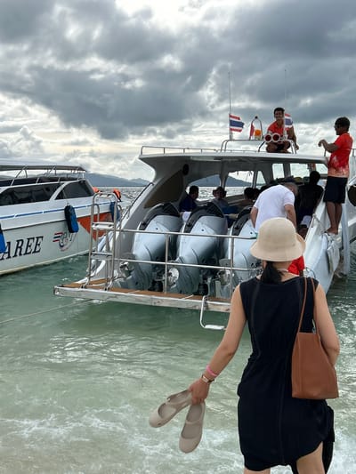 The image depicts a waterfront scene in Phru Nai, Thailand, with two speedboats moored near a pier. In the foreground, a woman wearing a black dress and a straw hat holds a pair of light-colored shoes. She is facing away from the camera, looking towards the boats.
The speedboat on the right has three outboard motors visible at the stern and several passengers inside. A man in an orange shirt, likely a crew member, is perched on the edge of the boat, holding what appears to be binoculars and smiling. Another man in a red shirt is standing near him, facing the water. Thai flags are attached to the boat's railing.
The speedboat on the left, named "PAREE" and possibly "PAREE PTY", is also visible. It has an orange stripe along its side and is docked with blue fenders. The water is choppy and a light green color, with some white foam near the boats. The sky is overcast with dramatic cloud formations, suggesting a change in weather. The background shows a hazy coastline with hills or mountains.