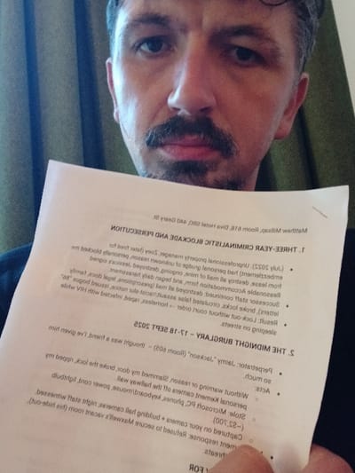 A man with dark hair, a mustache, and a goatee, wearing a dark shirt, is depicted in a chest-up selfie, looking directly at the camera. He holds a white document in his right hand, partially obscuring his chin and lower face. The document, oriented upside down, explicitly states "Matthew Milliam Room 818, Diva Hotel, 440 Geary St, San Francisco, United States." Further legible text details "1. THREE-YEAR CRIMINALISTIC AND BLOCKADE" and "2. THE MIDNIGHT BURGLARY - 17-18 SEPT 2022," listing "Perpetrator: Jaimy, 'Jackson' (Room 808)" and "Acts" including the theft of a Microsoft Kinect camera and iPad, a security response, a lock-out without a court order, and the subject "sleeping on the streets." The background consists of a vertically striped, olive-green curtain or fabric, indicative of an indoor setting, likely a hotel room.