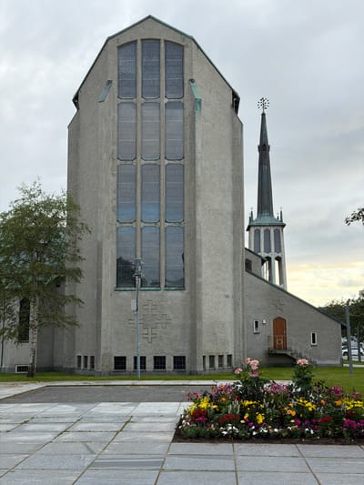 The image captures a view of Bodø Cathedral in Bodø, Norway, on an overcast day. The cathedral's main facade is a tall, grey structure with a steeply pitched roof. Dominating the facade is a series of tall, narrow stained-glass windows arranged in two vertical columns. Below these windows, a large, geometric cross-like relief is carved into the concrete. To the right of the main facade, the cathedral's slender, dark green spire rises against the cloudy sky, topped with a decorative element that resembles a starburst or a navigational buoy.
The foreground is paved with large, light grey square tiles arranged in a grid pattern, with some grass peeking through the gaps. A well-maintained flower bed is situated in front of the church building, showcasing a vibrant array of colorful flowers including pink dahlias, yellow marigolds, red poppies, and various other blooms.
To the left of the cathedral, a mature birch tree with sparse foliage stands, its trunk textured and light in color. The ground around the building is covered in green grass. The overall atmosphere is serene and somewhat muted due to the overcast sky. The architecture appears modern with clean lines and a focus on geometric shapes. The entrance to a smaller section of the church, located to the right of the main facade, is visible with a wooden door and a small set of stairs leading up to it.