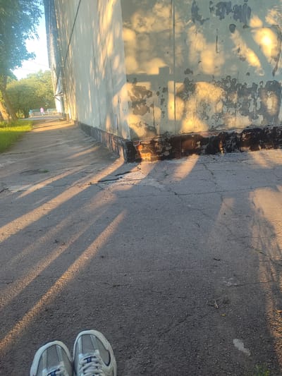 The image shows a perspective view of a paved path alongside a dilapidated building, with a person in the distance. The scene is illuminated by the warm, golden light of what appears to be late afternoon or early morning sun, casting long, distinct shadows of trees across the path and the building's wall.
The building's wall is a light, muted color, heavily weathered with peeling paint and patches of exposed brickwork. A dark, rough band runs along its base. The paved path is cracked and shows signs of wear, with occasional patches of grass growing through. To the left of the path, there is a strip of green grass and trees. In the far distance, a person is walking away from the camera, dressed in pink, on what appears to be another path or sidewalk adjacent to the building. The foreground shows the top of a pair of athletic shoes.
The overall atmosphere is serene and somewhat melancholic, due to the aging building and the long shadows. The location is identified as Zhovti Vody, Ukraine.