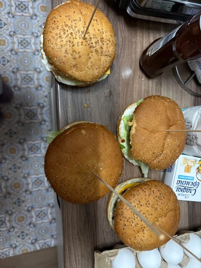 The image shows a tabletop arrangement of four hamburgers, each secured with a toothpick. A bottle of condiment and a carton of eggs are also visible on the table. The tabletop appears to be in a kitchen environment with a tiled floor. There is no evidence to suggest the location of this image is Rudartsi, Bulgaria. The description is based purely on the visual elements present in the image.