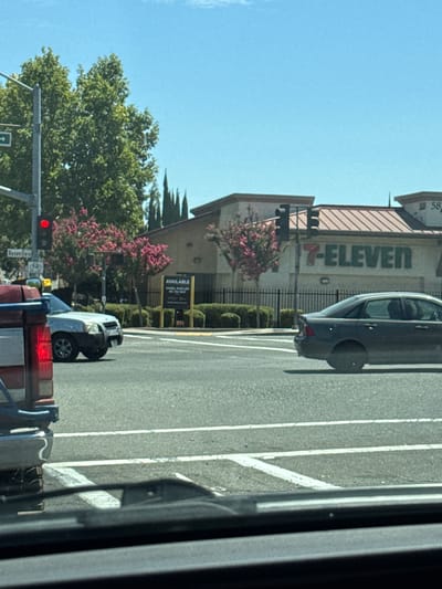 The image depicts a street scene in Foothill Farms, California, under a clear blue sky. A 7-Eleven store is visible in the background with its distinctive green and orange logo. Several vehicles are present, including a dark sedan driving on the road and a light-colored SUV stopped at an intersection. A red pickup truck is partially visible in the foreground, with its taillight illuminated. Traffic signals and street signs, including one for "Roseville," are also present. Lush green trees and flowering bushes add to the urban landscape. The scene appears to be captured from a vehicle, indicated by the presence of what looks like a dashboard or windshield in the lower part of the frame.