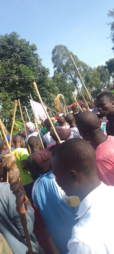The image shows a group of people holding long wooden sticks, some with a white cloth attached. The crowd is dense, and individuals are packed close together, mostly obscured from view. They are standing on a dusty dirt road with trees and power lines above them. The day is clear and sunny. The scene is likely an outdoor gathering in Isukha, ICHINA, Kenya, based on provided coordinates. The atmosphere is informal, with people interacting casually as part of a larger gathering.