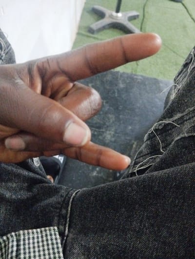 The image shows a close-up of a person's hand, with dark skin, making a "rock on" hand gesture. The hand is positioned in the foreground, with fingers extended and the thumb tucked in. The person is wearing dark-colored denim jeans, visible at the bottom and right of the frame. The texture of the denim is rough, with some loose threads. The background is blurred, showing a hint of a green surface and what appears to be a chair base. The lighting suggests an indoor setting, possibly during the daytime. There is no visible text in the image. The provided location context is Barikin Gidar Kanat, Nigeria.