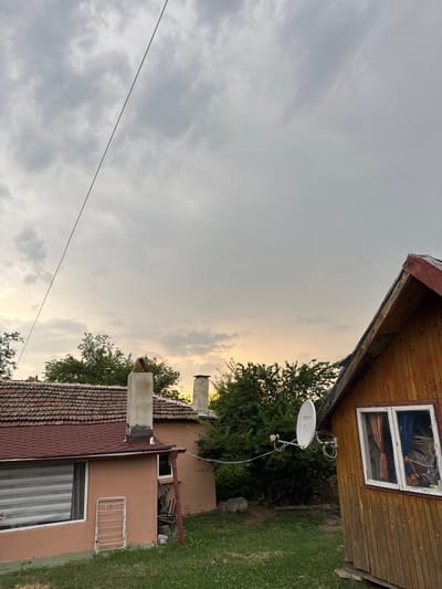 The image depicts a view of residential buildings and the sky in Sredets, Bulgaria. In the foreground, a wooden structure with a slanted roof and a window is visible on the right. To the left, a smaller building with a tiled roof and chimneys stands. A satellite dish is mounted on the exterior wall of the wooden structure, and trees and a grassy area surround the buildings. The sky above is overcast with clouds, and the light suggests it may be late afternoon or early evening. There are no people or discernible actions taking place.