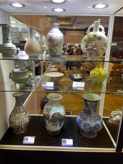 The image displays a collection of antique porcelain vases exhibited in a glass display case. The vases vary in size, shape, and decoration, featuring intricate patterns, floral motifs, and scenic landscapes. Some vases are adorned with Chinese calligraphy. A label with Chinese text is visible on one of the display shelves. The setting appears to be an exhibition or a museum, possibly in Futian District, China, given the location context. The lighting within the display case highlights the artifacts. The overall atmosphere suggests a cultural display of fine art and historical craftsmanship. No people are clearly visible within the display, but reflections in the glass indicate the presence of visitors or staff in the background. The image is oriented vertically, presenting a close-up view of the items.
