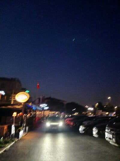 The image shows a street scene at night in Istanbul, Turkey, specifically at the coordinates 40.956 latitude and 28.821 longitude. The scene is dark, with a few streetlights illuminating the road. There are several cars parked on both sides of the road. A few individuals are walking on the sidewalk in the distance. The camera appears to be positioned inside a vehicle, facing forward toward the street. The street is paved and has streetlights on either side. There is a Turkish flag on a pole in the foreground, and a large orange sign that is illegible. The sky is dark blue with two small bright lights. The image is taken from a street in Istanbul, Turkey. The cars are parked and illuminated by streetlights. It is evening, with a clear dark sky, and the road is paved. The image is blurred, suggesting it was taken from a moving vehicle.