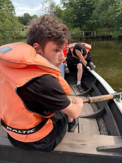 In the city of Allerslev, Denmark, two individuals are seated in a wooden rowboat on a body of water. The individual in the foreground, facing away from the camera, wears an orange life vest over a black t-shirt and black shorts, with the brand "BALTIC" visible on the vest. This person's hands are gripping an oar, positioned for rowing. The second individual, further back in the boat and partially obscured, also wears an orange life vest and a dark hooded garment, with their face covered by their hand. The environment features a tranquil body of water, with lush green trees and vegetation lining the banks. In the background, a wooden bridge or walkway is visible extending over the water. The sky above is partly cloudy.
