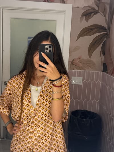 A young woman with long, dark hair is taking a mirror selfie in what appears to be a bathroom. She is wearing a white tank top under a collared, button-up shirt with a geometric print in shades of pink, yellow, and black. She has several necklaces and bracelets on, and a ring on her left hand. She is holding a black smartphone up to take the picture. The background shows a wall with floral wallpaper, a light switch panel, and a black trash can. A door is visible to the left of the woman. The image is taken indoors, in a residential or public restroom setting. There is no visible indication of the location being in Boca Raton, United States of America.