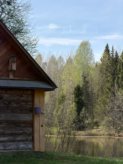 The image shows a rustic wooden building with a steeply pitched roof covered in corrugated metal. A small birdhouse is attached to the side of the building near the roofline. Adjacent to the building, there is a view of a lake or pond surrounded by trees, including evergreens and deciduous trees with budding leaves. The sky is partially visible and appears to be lightly cloudy. The scene appears to be rural and wooded. The specific location is Ural'skoe, Russia. There is no indication of people or interaction shown in the photo.