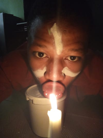 A close-up photograph shows a person with dark skin and tribal-style white markings on their forehead and around their nose. Their eyes are dark brown, and their mouth is open with their tongue extended. A lit white candle in a white container is directly in front of their mouth, appearing as if they are about to extinguish the flame. The background is dark, but a piece of furniture is visible in the top left. The location, Oshogbo, Nigeria is not discernible within the frame.