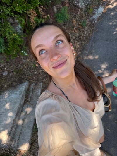 A young woman with striking blue eyes looks up and to her left, a slight smile on her lips. She is wearing a light beige blouse with a gathered neckline and a thin silver necklace. Her brown hair is pulled back into a ponytail. She appears to be outdoors, with a backdrop of green foliage and dry ground. To her right, a brown purse is visible, and she is holding a green bottle. The lighting suggests it is daytime, with shadows indicating the sun's position. The scene takes place on a paved path, with some concrete steps to the left.
