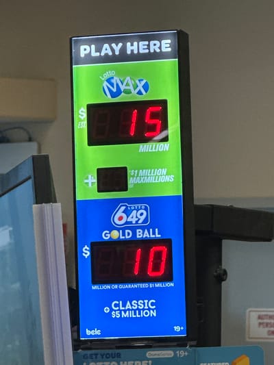 This is a close-up, eye-level, vertical shot of a digital lottery display in Coquitlam, Canada. The display shows two lottery games: Lotto Max and Lotto 6/49 Gold Ball. The top section, dedicated to Lotto Max, features the logo and a jackpot amount displayed as "$15 MILLION". Below this, it indicates "$1 MILLION MAXMILLIONS". The bottom section, for Lotto 6/49 Gold Ball, shows a jackpot of "$10". It also mentions "MILLION OR GUARANTEED $1 MILLION" and "CLASSIC $5 MILLION". The BCLC logo is visible at the bottom of this section. The display is lit and the numbers are bright red. The background is a neutral gray, likely a wall or other surface in a retail setting. There are no people or animals visible in the frame.