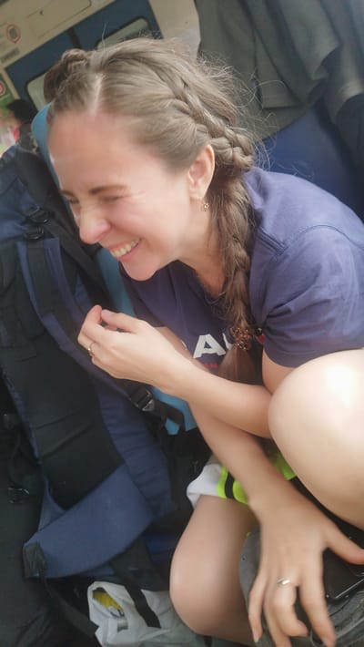 A woman with braided brown hair laughs heartily, her eyes squeezed shut. She is wearing a dark blue t-shirt with a white logo on it, and a gold earring. She is sitting with her knees pulled up to her chest, next to a large navy blue backpack. The background appears to be the interior of a train or bus, with blue seats and windows visible. The lighting suggests it is daytime. The woman's laughter and relaxed posture convey a sense of joy and amusement. The visible text on her shirt appears to be "A".