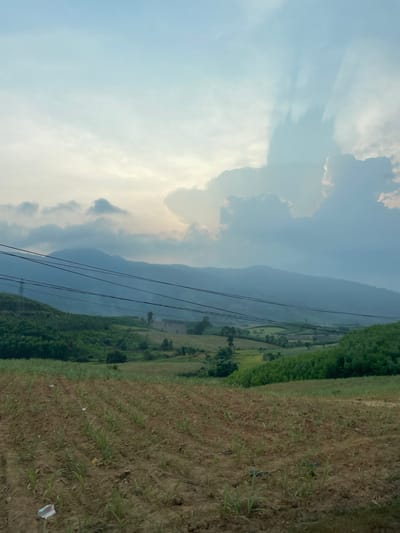 The image shows a landscape in Thôn 18, Vietnam. The sky is partially cloudy, with light blue patches and large, billowing clouds. A mountain range fades into the distance, obscured by haze or mist. Power lines stretch across the scene, leading towards the mountains. Patches of green fields and trees are interspersed among areas of cultivated land. In the foreground, there is a field with dry grass and sparse vegetation. Some scattered debris, possibly plastic or paper, is visible on the ground. The image is taken at an angle, with the camera likely tilted to the side.
