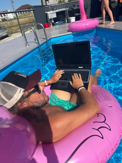 The image shows a person lying on a pink inflatable raft in a swimming pool, using a laptop. The person is wearing swim trunks, sunglasses, and a baseball cap. Their legs are partially submerged in the blue water. A pink inflatable ring is also visible in the pool. The scene appears to be outdoors on a sunny day, likely in a residential or resort setting, given the presence of a swimming pool and surrounding deck. The text on the inflatable raft appears to be a warning label, with some portions unreadable due to the image quality and angle. The overall mood seems relaxed and leisurely, with the person multitasking by working on their laptop while enjoying the pool.