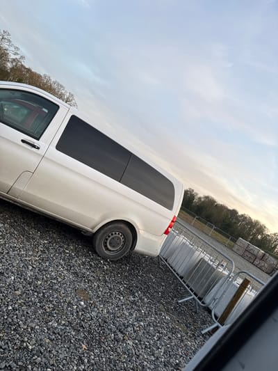 A white panel van, resembling a Mercedes-Benz Vito, is parked on a dark grey gravel surface at Baldurgan Hill, Ireland. The vehicle is observed from its left side, displaying clear front side windows with a reddish interior component visible, and tinted black rear side windows. The lower body panels exhibit dirt accumulation, and the rear left wheel is a steel rim without a hubcap. To the right of the van, a series of silver metal crowd control barriers are positioned, with some supporting or resting against large, light-colored concrete blocks. The background features an unpaved area, a distant treeline of bare deciduous trees, and stacks of building materials (possibly concrete slabs) on the far right. The sky overhead is overcast with light blue and grey hues. No individuals, actions, or direct interactions are depicted in the scene.