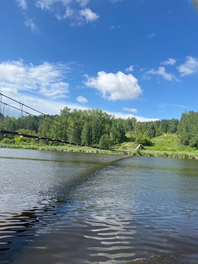 A daytime, low-angle shot captures a view across a wide, calm river towards a lush green, tree-covered bank. A long, wooden suspension bridge spans the river, its weathered planks and metal cables visible. The bridge extends from the left side of the frame and curves slightly towards the right bank, where it appears to connect to a wooden structure on a grassy hill. The water in the foreground is dark and reflective, with ripples disturbing its surface. The sky above is a vibrant blue, dotted with fluffy white clouds. The scene depicts a natural landscape, likely in a rural or park setting. The location is the town of Kamensk-Uralsky, Russia, specifically the suspension bridge over the Iset River.