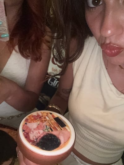 The image is a close-up, tilted shot of a person lying down, with their face and upper body visible. The person has dark hair, an open eye with dark eyelashes, and glossy, pink lipstick. They are wearing a light-colored, ribbed tank top. In the foreground, held by the person's hand, is a round object with an image of Gustav Klimt's "The Kiss" painted on it. The background shows a blurred scene with what appears to be a light-colored blanket or bedding. Some text is partially visible on a patterned fabric behind the person, including the word "Est." and "Gir". The image has a warm, intimate feel, possibly taken indoors during the day or evening. The location context provided is Valletta, Malta.