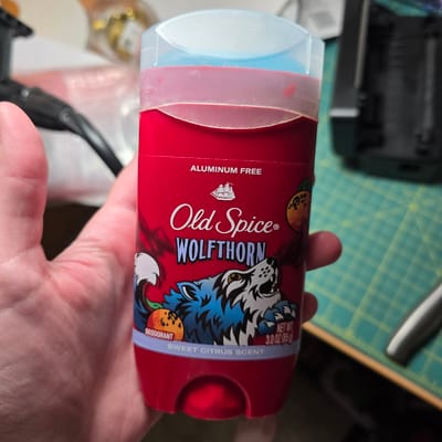 A hand holds an Old Spice Wolfthorn deodorant stick. The deodorant stick is red with a blue and white wolf graphic. The text on the stick reads "ALUMINUM FREE", "Old Spice", "WOLFTHORN", "DEODORANT", "NET WT. 3.0 OZ (85g)", and "SWEET CITRUS SCENT". The background is blurred, but appears to be a desk with a cutting mat and other objects. The image is not geotagged and the location in Portland, United States of America is not specified.