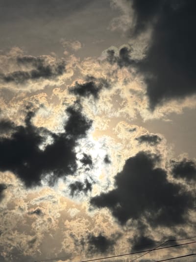 The image depicts a dramatic sky filled with dark, billowing clouds illuminated by a bright, partially obscured sun. The sunlight filters through the clouds, creating a contrast between light and shadow and casting an almost ethereal glow on some of the cloud formations. In the lower portion of the image, a series of utility wires are visible, stretching horizontally across the frame, suggesting an urban or semi-urban setting. The overall mood is somber yet awe-inspiring, characteristic of a turbulent weather system. There are no people or specific structures visible in this image. The location is identified as Abovyan, Armenia.