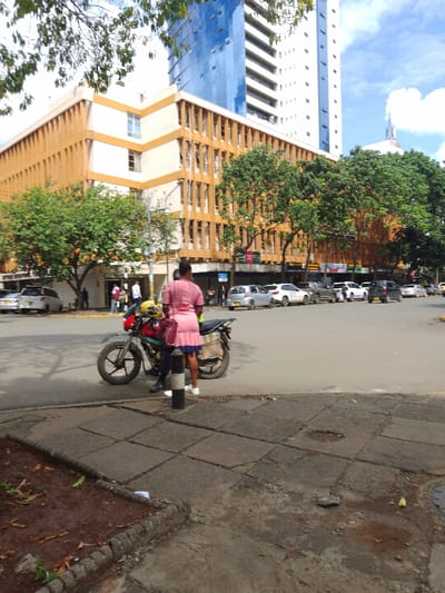 The image shows a street corner in Athi River, Kenya, at the intersection of a paved road and a sidewalk. In the foreground, there is a parked motorcycle with a woman standing next to it. She is wearing a pink dress and carrying a pink purse. She is speaking to a man who is wearing a yellow helmet and a black jacket. The man is holding a yellow helmet. The sidewalk is made of large concrete slabs. In the background, there is a street lined with parked vehicles. The street is busy with cars and pedestrians. The street is lined with trees and buildings, including a tall, modern office building. The building is painted blue and has many windows. It is located on the left side of the image. There are several other buildings in the background, including a yellow building with a large sign. The sky is clear and blue with a few clouds. The scene is situated in Athi River, Kenya, at the intersection of a paved road and a sidewalk.