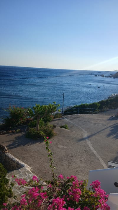 This media file depicts a serene coastal scene in Kerames, Greece, likely during the daytime, as indicated by the bright, clear sky and the way the sun casts shadows. The primary subjects are the vast expanse of the deep blue sea meeting the bright blue sky, a paved pathway, and vibrant pink bougainvillea flowers cascading down a wall.
The scene is set outdoors, alongside the sea. The pathway appears to be a pedestrian walkway or a narrow road, bordered by a low blue railing on one side, separating it from the sea. The sea itself is calm, with gentle ripples and small, dark shapes that might be rocks or buoys visible in the distance. Lush green vegetation, including small trees and bushes, grows along the edge of the pathway and the sea. The most striking element is the profusion of bright pink bougainvillea flowers, their colors contrasting beautifully with the blue of the sea and sky, and the gray of the pathway and stonework.
There are no people or animals visible in the frame, contributing to the peaceful and tranquil atmosphere. The stonework along the lower edge of the pathway suggests a terraced or elevated position, offering a vantage point over the sea. The overall impression is one of natural beauty and a peaceful, sunny day in a Mediterranean coastal location. No visible text is present in the image.